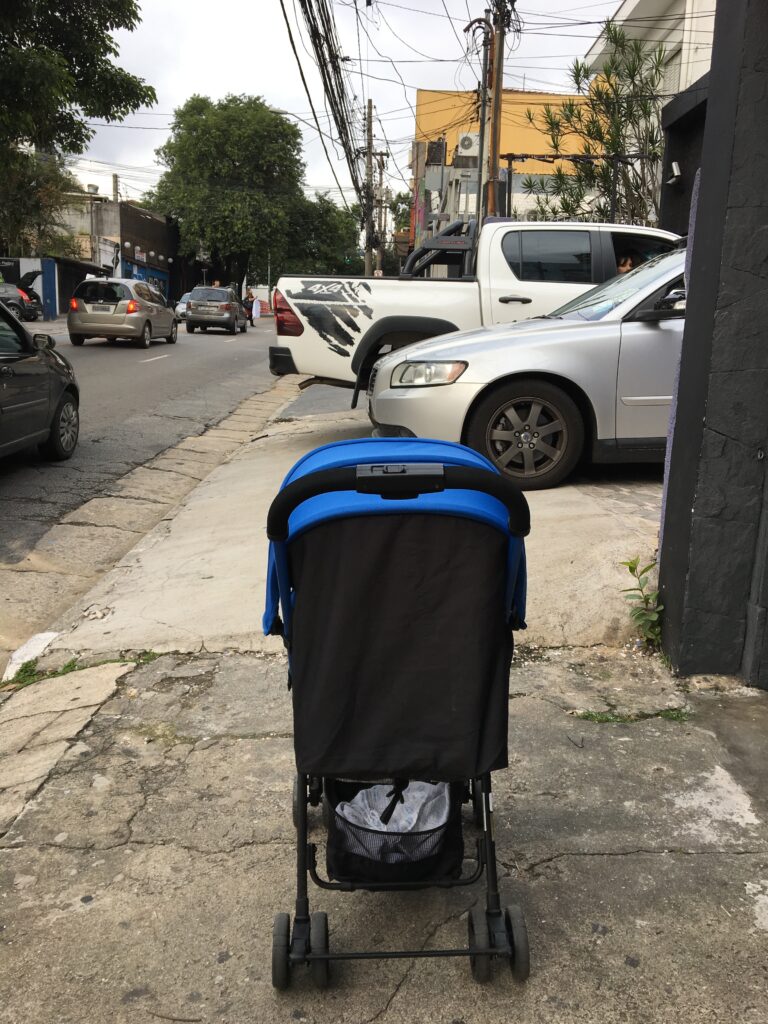 Um carrinho de bebê azul na calçada, carros estacionados bloqueando a passagem
